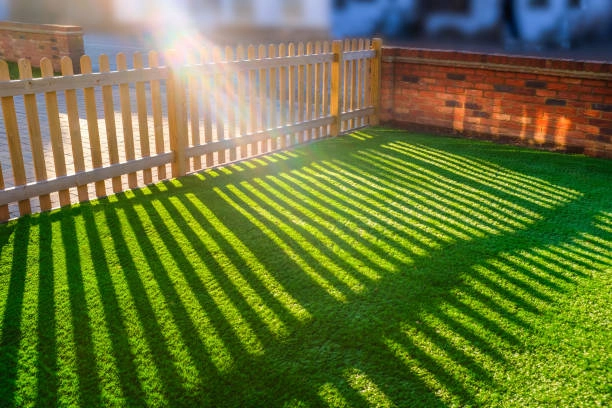 Sunlight casting fence shadows on green grass beside brick wall. -artificial turf grass